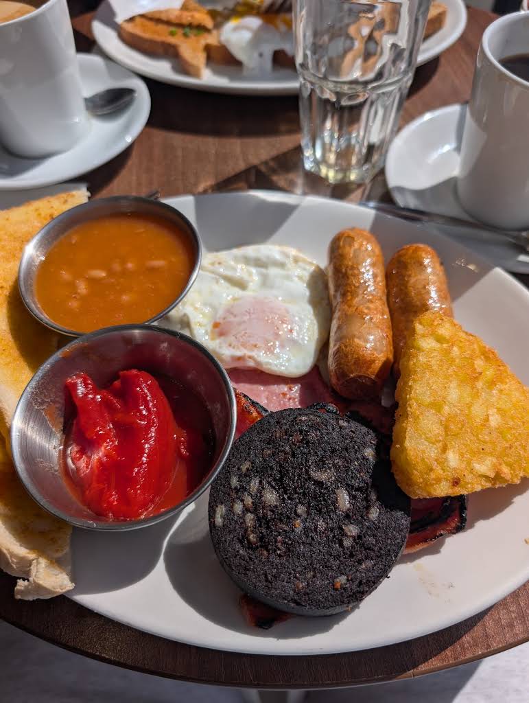 Breakfast at Café 24, with eggs, sausages, bacon, beans, tomatoes and toast on a sunny wooden table.