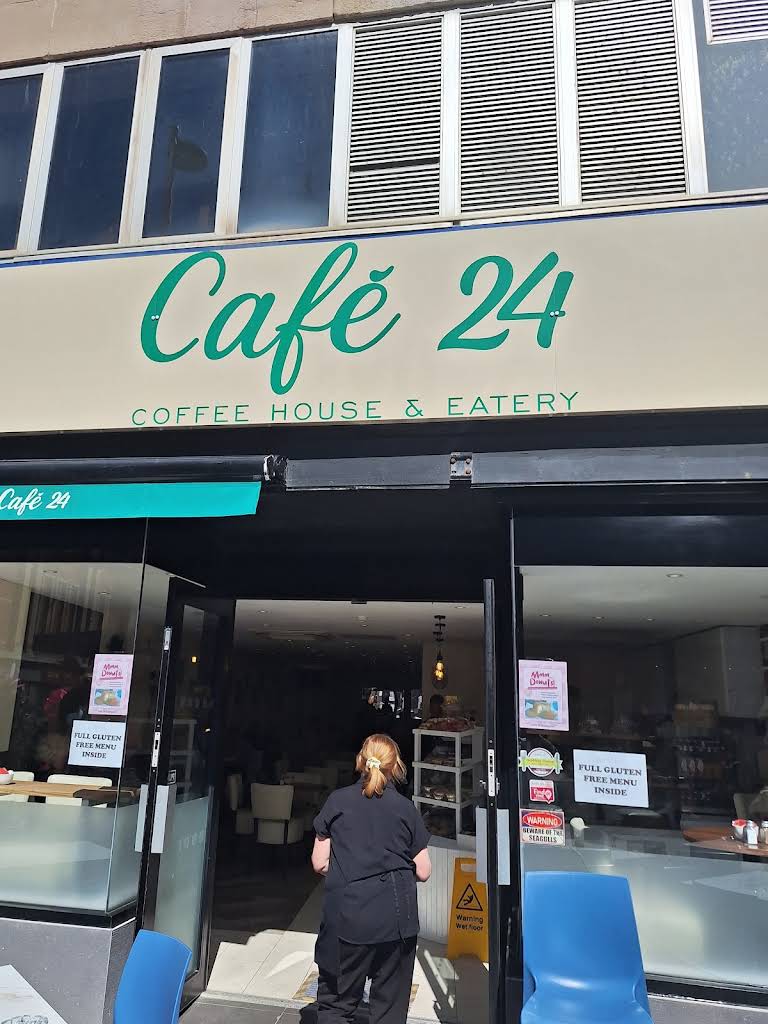 Café 24’s complete green script sign on a cream board, black-framed entrance and visible doorstep.